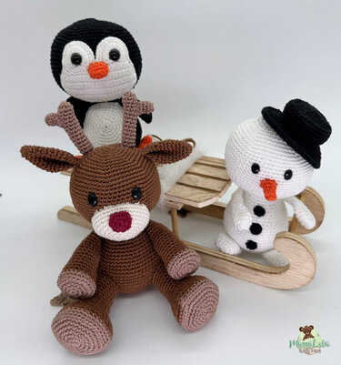 Gehäkeltes Rentier, Pinguin und Schneemann mit schwarzem Hut neben einem kleinen Holzschlitten