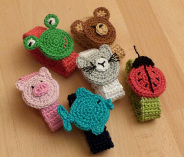 Sechs gehäkelte Kinderarmbänder mit Tierköpfen (Frosch, Bär, Katze, Schwein, Fisch, Marienkäfer) auf hellem Holz