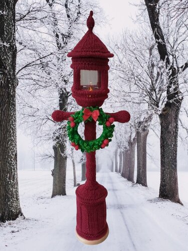 Maroon knitted lantern cover decorated with a green wreath and lit candle, set on a snowy tree-lined path