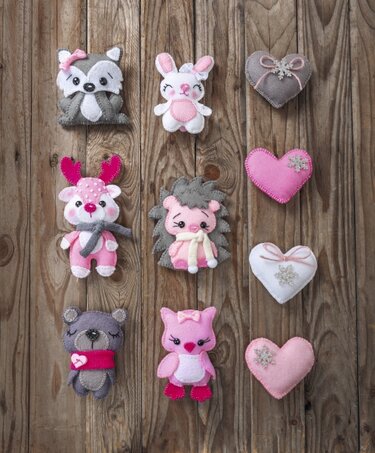 Genähte Filztiere und Filzherzen in Rosa, Grau und Weiß auf Holzbrett, darunter Fuchs, Hase, Igel, Bär, Eule und Rentier.