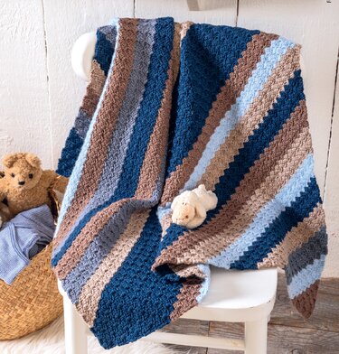 Gehäkelte Babydecke mit texturierter Maschenstruktur und breiten Streifen in Blau-, Grau- und Brauntönen, drapiert auf weißem Stuhl mit kleinem Stofftier