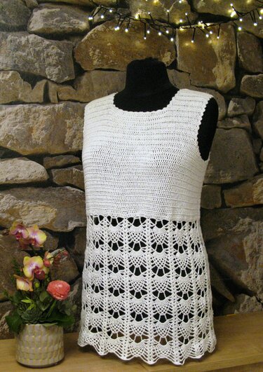 Weißes ärmelloses Häkeltop mit Muschel‑Spitzenmuster und Rundhalsausschnitt auf schwarzem Mannequin vor Steinwand und Lichterkette.