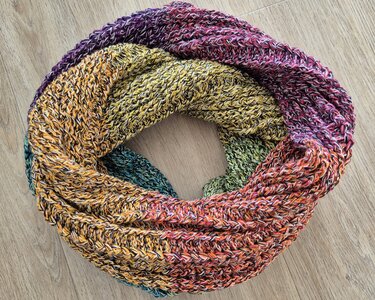 Gehäkelter Loop mit verlaufendem Garn in Gelb, Grün, Orange, Rot und Violett auf Holzuntergrund