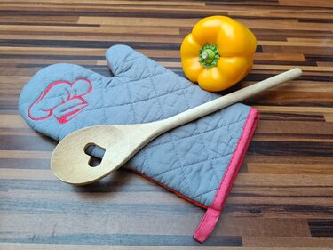 Grau gesteppter Topflappen mit roter Kochmützen-Stickerei, Holzlöffel mit Herzausschnitt und gelbe Paprika auf Holzarbeitsfläche