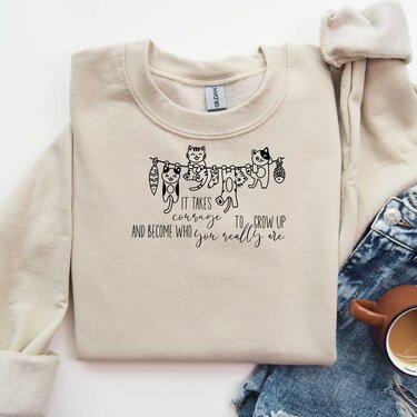 Hellfarbener Pullover mit schlichtem schwarzen Plottdesign: mehrere sitzende und hängende Katzen über einem englischen Zitat.