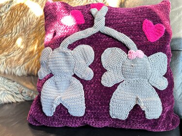 Dunkelviolettes Häkelkissen mit zwei grauen Elefanten, deren Rüssel ein Herz bilden, sowie einem kleinen rosa Herz.