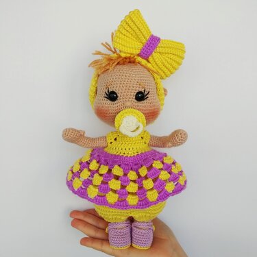 Amigurumi crochet baby doll with a large yellow bow, pacifier, and purple-and-yellow dress, shown in a hand for scale.