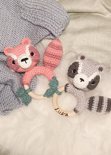 Rosa gehäkelter Fuchs- und grauer Waschbär-Greifling mit Holzringen auf weicher Decke