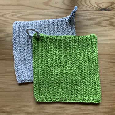 Zwei gestrickte Topflappen, einer hellgrau und einer lindgrün, mit Aufhängeschlaufen auf einem Holztisch.