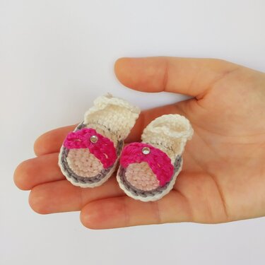 Pair of tiny crocheted sandals with pink straps resting on a human palm to show scale