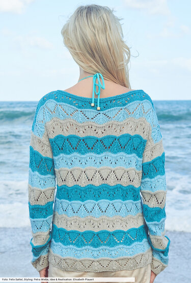 Rückenansicht einer Frau am Strand in gestreiftem Strickpullover in Blau- und Beigetönen mit Ajourmuster.