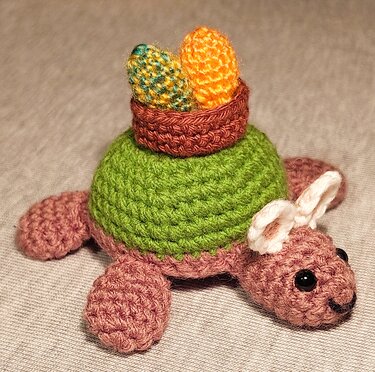 Gehäkelte Amigurumi-Schildkröte mit weißen Hasenohren und Körbchen mit zwei Ostereiern auf dem grünen Panzer