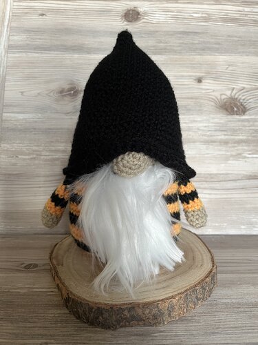 Gehäkelter Wichtel mit schwarzem Spitzhut, weißem Bart und orange‑schwarzen Streifen, stehend auf einer Holzscheibe.