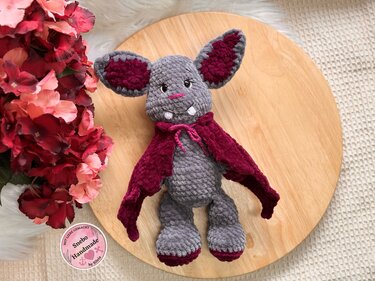 Grau-bordeauxfarbene gehäkelte Amigurumi-Fledermaus mit Umhang auf runder Holzplatte