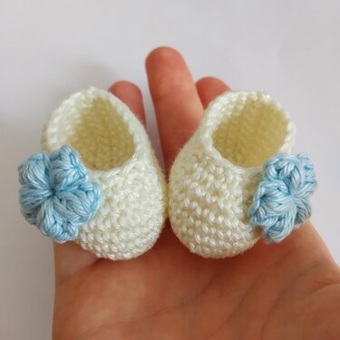 Winzige gehäkelte Babyschühchen in Creme mit hellblauer Blume auf einer Handfläche