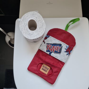 Rote genähte Kosmetiktasche mit Pop‑Art 'YEAH!'-Einsatz, ovaler Entnahmeschlitz und grüner Aufhängeschlaufe neben einer Toilettenpapierrolle auf weißem Hintergrund