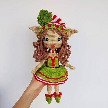Hand holding a crocheted amigurumi Christmas elf doll with curly brown hair, a green dress, striped hat, and curled-toe shoes.