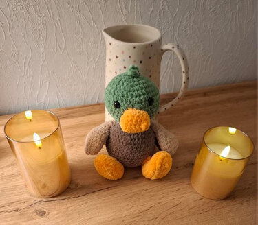 Gehäkelte Amigurumi-Ente mit grünem Kopf und braunem Körper zwischen zwei warmen LED-Kerzen auf einem Holztisch.