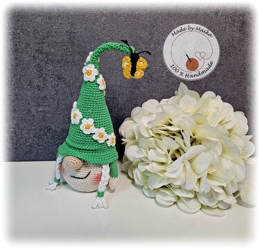 Amigurumi-Wichtel mit grünem Zipfelhut, weißen Gänseblümchen und kleinem gelben Schmetterling, neben einer großen weißen Blüte.