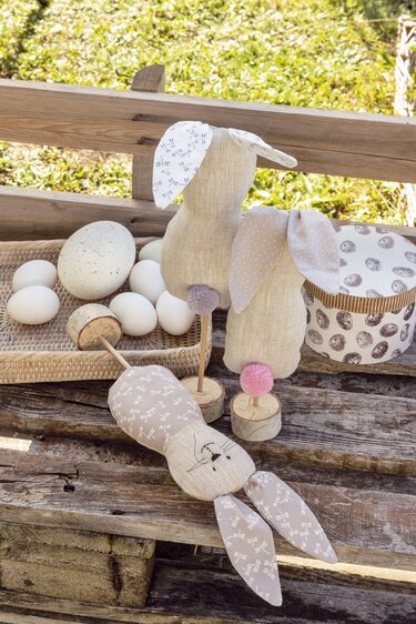 Drei genähte Stoffhasen, zwei auf Holzständern, mit Pompon‑Schwänzen neben Dekoeiern auf rustikalem Holz