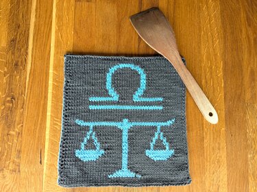 Gestrickter quadratischer Topflappen in Dunkelgrau mit türkisfarbener Waage, auf Holztisch neben Holzspatel.