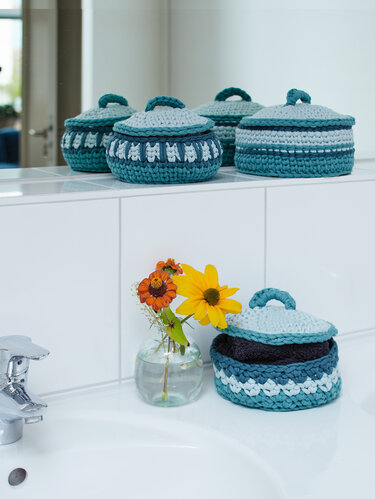 Gehäkelte Körbe mit Deckel in Blau- und Türkistönen, gefüllt mit Handtüchern neben einer Vase auf der Badablage