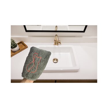 Graues Handtuch mit roter, stilisierter Katzenstickerei, von einer Hand über einem Waschbecken gehalten.