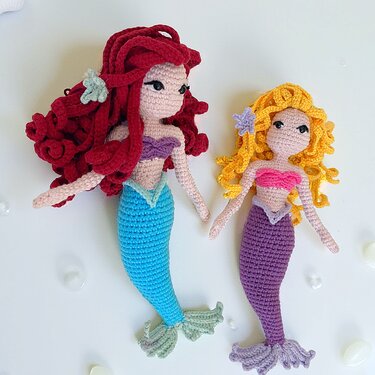 Zwei Amigurumi-Meerjungfrauenpuppen nebeneinander, eine mit roten Locken und türkisem Schwanz, die andere mit gelben Locken und violettem Schwanz