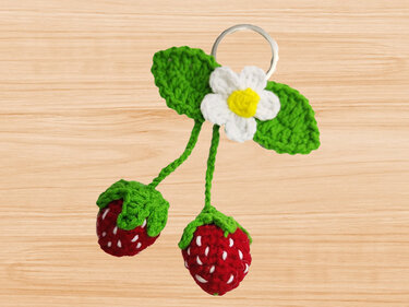 Crocheted strawberry keychain with two red berries, green leaves and a white flower on a metal ring on a light wood background