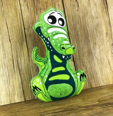 Grünes Krokodil-Kuscheltier mit großen Augen und weißen Punkten, sitzend vor Holzpaneelen.