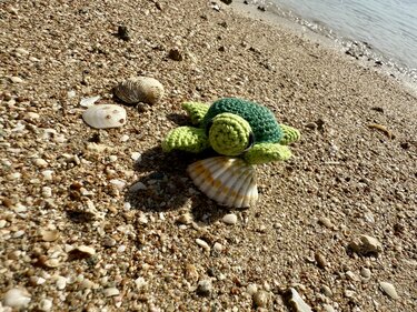 Gehäkelte grüne Meeresschildkröte (Amigurumi) auf Muschel am Strand.
