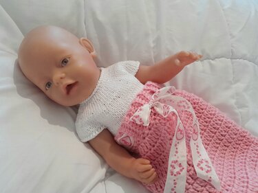 Babypuppe liegt auf weißer Decke, trägt weißes gestricktes Oberteil und rosa gehäkelten Rock mit Band