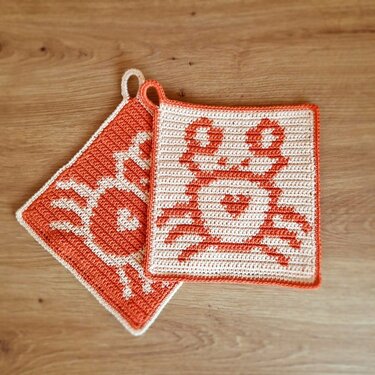 Zwei gehäkelte quadratische Topflappen in Orange und Creme mit Krabbenmotiv auf Holzuntergrund