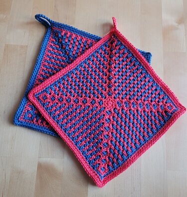 Zwei quadratische gehäkelte Topflappen in Blau und Pink mit diagonalem Muster und Aufhängeschlaufen auf hellem Holzhintergrund.