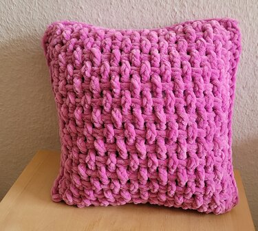 Quadratisches gehäkeltes Kissen in intensivem Fuchsia mit grober, kuscheliger Maschenstruktur auf hellem Holz