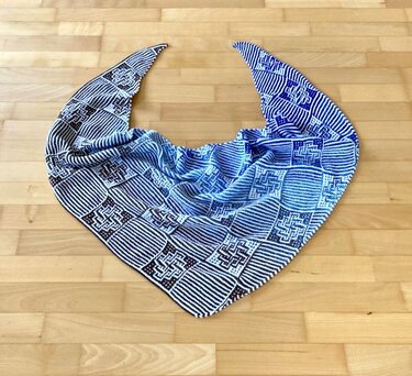 Gestricktes Dreieckstuch in Blau und Weiß mit Streifen- und Quadratmustern auf hellem Holzboden.