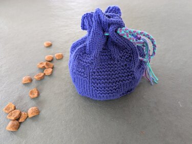 Lila gestrickter runder Leckerlibeutel mit geflochtener Kordel neben verstreuten Trockenfutterstücken