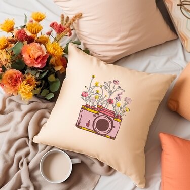 Beiges Kissen mit aufgestickter rosa Kamera und bunten Blumen, daneben ein Blumenstrauß und eine Tasse.