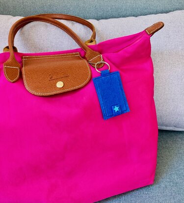 Leuchtend pinke Tasche mit braunen Tragegriffen, Lasche und blauem Filzanhänger mit hellblauem Stern auf hellem Kissenhintergrund.