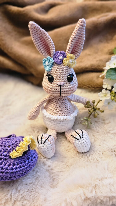 Gehäkelter Amigurumi-Hase in Pastellfarben mit drei bunten Blüten am Kopf und langen Ohren, sitzend auf weichem Untergrund