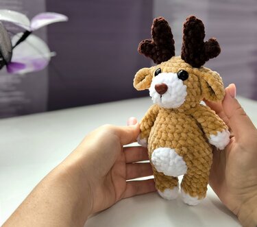 Hand hält gehäkeltes Rentier-Amigurumi in Beige mit weißem Bauch, Pfotenakzenten und dunkelbraunem Geweih