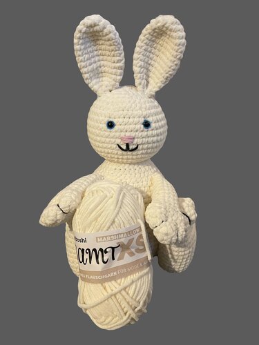 Weißer gehäkelter Amigurumi-Hase mit blauen Augen und rosa Nase, der ein cremefarbenes Garnknäuel hält.