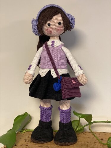 Hand-crocheted amigurumi doll in lavender vest, earmuffs, black skirt and boots, carrying a small purple crossbody bag.
