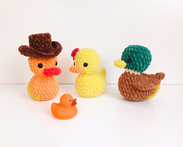 Three crocheted plush ducks (cowboy hat, yellow with red bow, mallard-colored) with a small orange rubber duck on a white background.