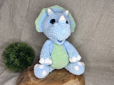 Blau-grüner gehäkelter Triceratops-Amigurumi mit weißen Hörnern und Punkten, sitzend auf einem Holzstück vor hellem Stoffhintergrund.