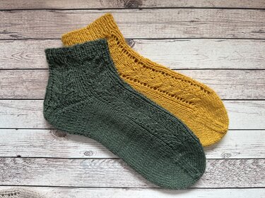 Zwei gestrickte Knöchelsocken auf hellem Holz, eine dunkelgrün mit Strukturmuster, eine senfgelb mit Lochmuster.