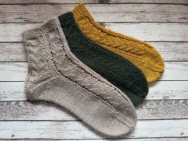 Drei gestrickte knöchelhohe Damensocken in Beige, Dunkelgrün und Senfgelb mit feinem Ajourmuster, nebeneinander auf hellem Holzhintergrund.