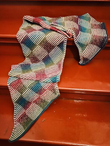 Mehrfarbiger gestrickter Schal mit diagonalen Karofeldern und weißen Streifen auf rotbraunen Treppenstufen.