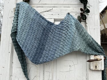 Gehäkeltes Dreieckstuch mit blau-grünem Farbverlauf und Muschelmuster, über einer verwitterten weißen Holztür drapiert