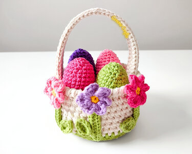 Beige crochet Easter basket with a handle, flower appliqués, and several pink, purple, and green crocheted eggs.